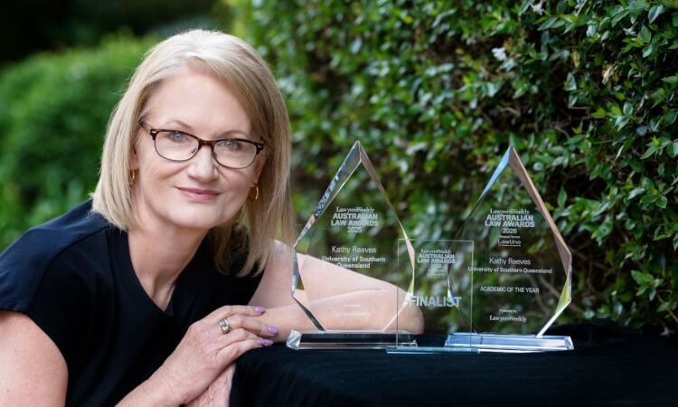 A woman with glasses smiles beside two clear glass awards placed on a black cloth, with greenery in the background.