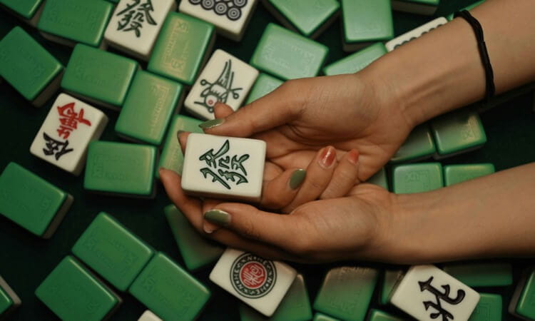A pair of hands holds a white mahjong tile with green characters above a pile of scattered green-backed mahjong tiles.