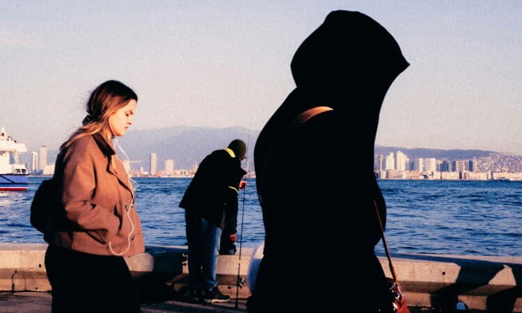 Three people stand near a waterfront; one wears a hood, one walks with earphones, and one fishes. City buildings and mountains appear in the background.