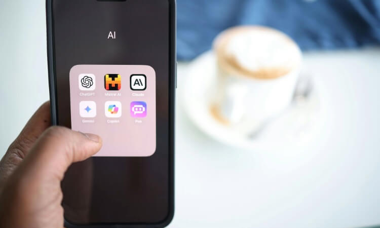 A hand holds a smartphone displaying a folder labeled “AI” with various artificial intelligence app icons, next to a blurred coffee cup.