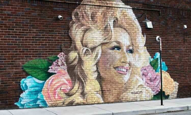 Mural of a smiling woman with blonde hair surrounded by colorful flowers painted on a brick wall along a sidewalk.