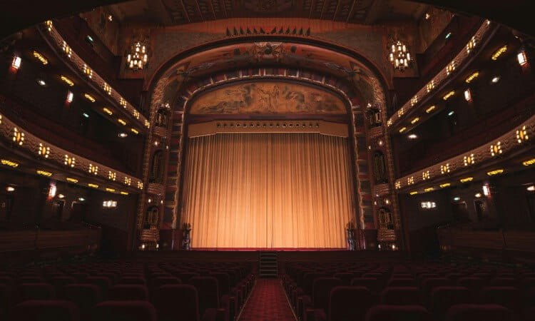 Empty theater auditorium with rows of red seats facing a large stage framed by ornate architecture and a closed gold curtain.
