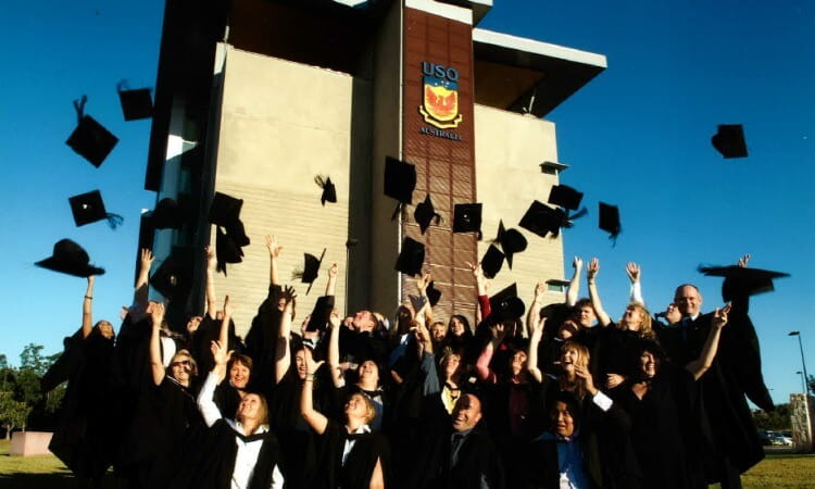 A group of graduates in caps and gowns toss their hats into the air in front of a university building on a sunny day.