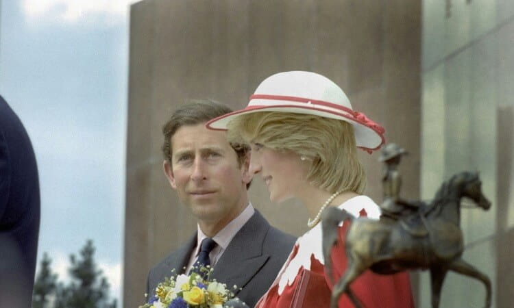 A man in a suit and a woman in a red and white outfit with a hat stand outdoors near a statue of a person on horseback; the woman holds a bouquet of yellow flowers.
