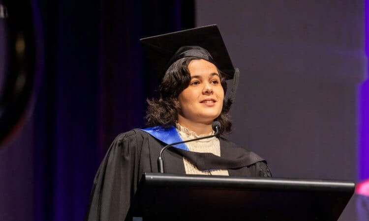 A person in a graduation cap and gown speaks at a podium during a ceremony.