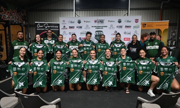 A women’s sports team poses indoors, each player holding a green jersey with white stripes. A banner with various sponsor logos is displayed in the background.
