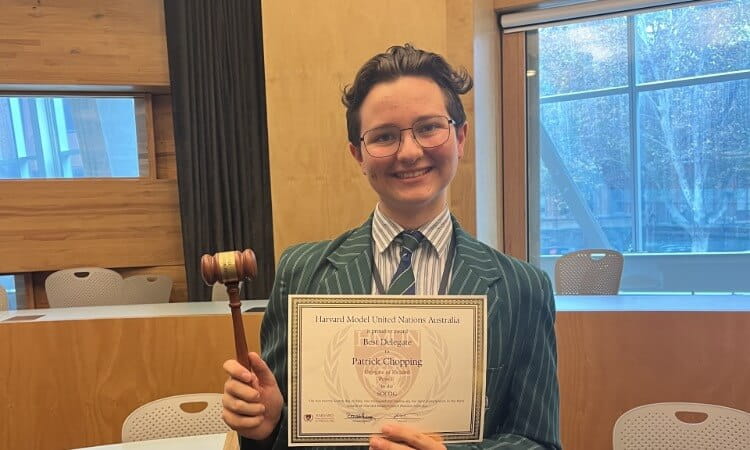 Person in a striped blazer holding a gavel and a certificate stands in a classroom with wooden walls and large windows.