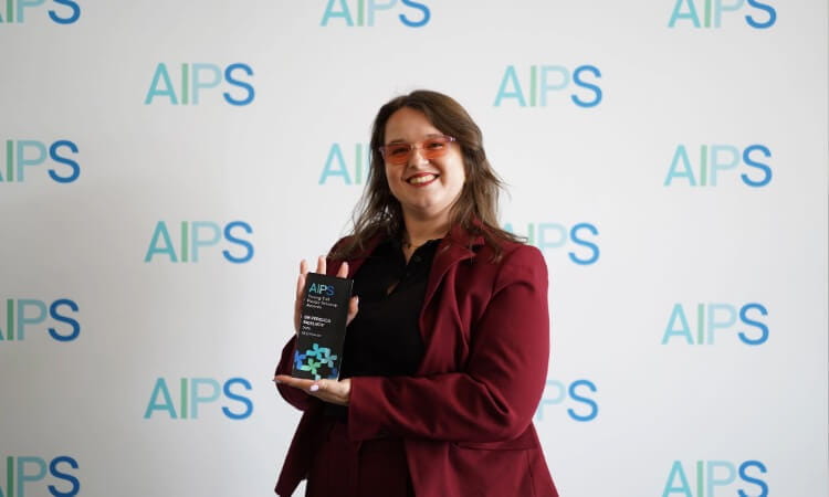 Person in a maroon suit holds an AIPS award trophy and smiles in front of a backdrop with repeated “AIPS” logos.