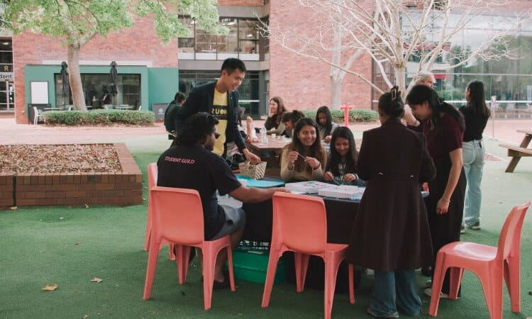 A group of people sit and stand around a table outdoors, talking and engaging in activities, with buildings and trees in the background.