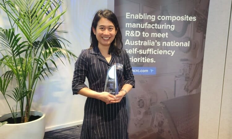 Woman holding an award stands in front of a banner about composite manufacturing R&D and Australia’s self-sufficiency priorities.