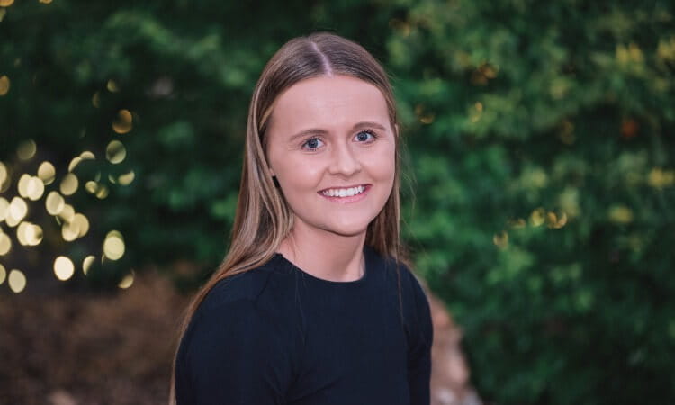 A young woman with straight light brown hair smiles at the camera, wearing a black top, with greenery and blurred lights in the background.