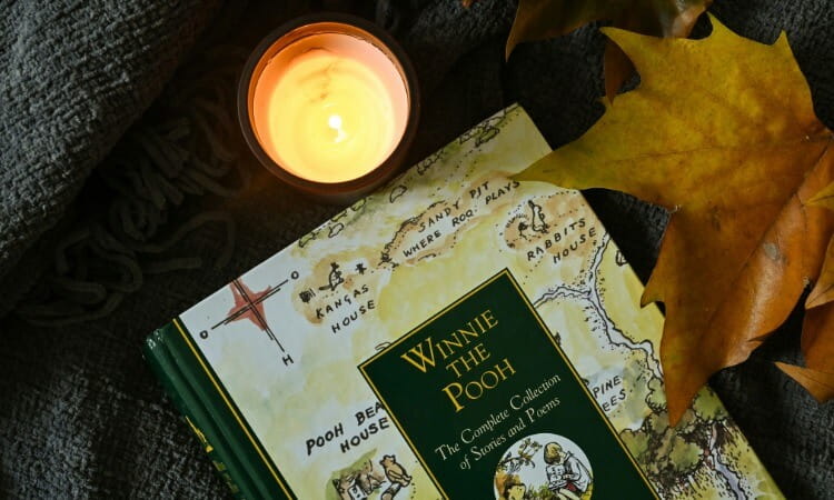 A copy of Winnie the Pooh: The Complete Collection of Stories and Poems resting near a lit candle and autumn leaves.