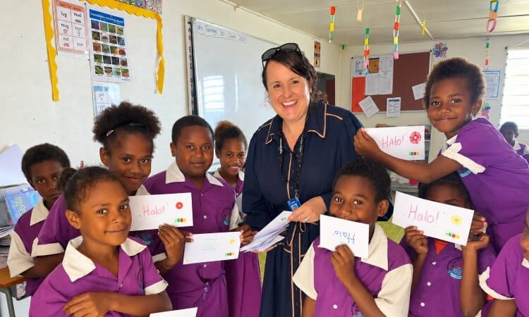 A teacher stands with a group of primary school students in purple uniforms who are holding handmade cards labeled “Halo!”.