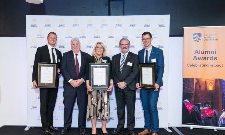 Five people standing in a row on a stage holding framed awards in front of a University of Southern Queensland branded backdrop.