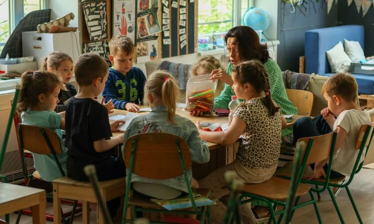 A teacher sits at a round table with a group of children in a classroom as they work on an activity.