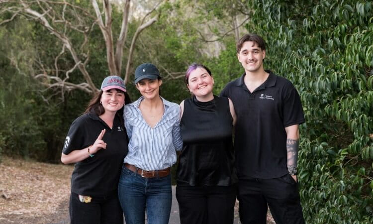Four people standing together outdoors in front of trees and greenery.