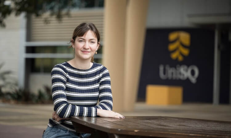 A young woman sitting at an outdoor table with a UniSQ logo visible in the background.