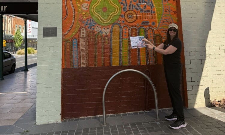 A person in a black shirt and cap stands next to a patterned brick mural, pointing at a small flyer held in their hand.