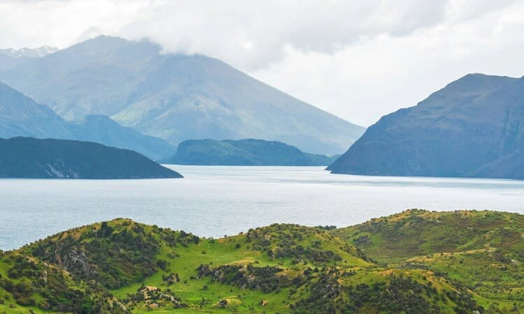 View of a large lake surrounded by rolling green hills and distant mountain ranges under a cloudy sky.