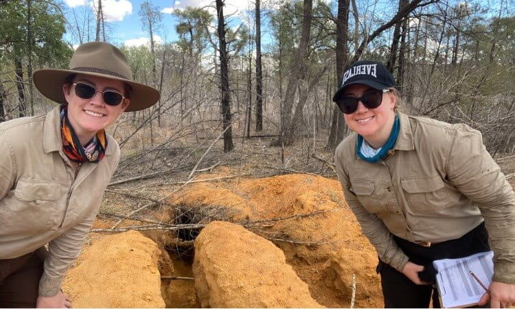 Two people in field clothes and sunglasses crouch by a large hole in red dirt in a forest.