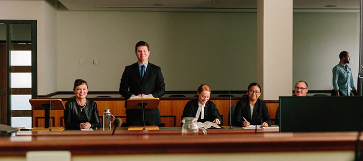 Five individuals in a courtroom setting, with one standing at the podium and the others seated at a table, appearing focused and prepared.