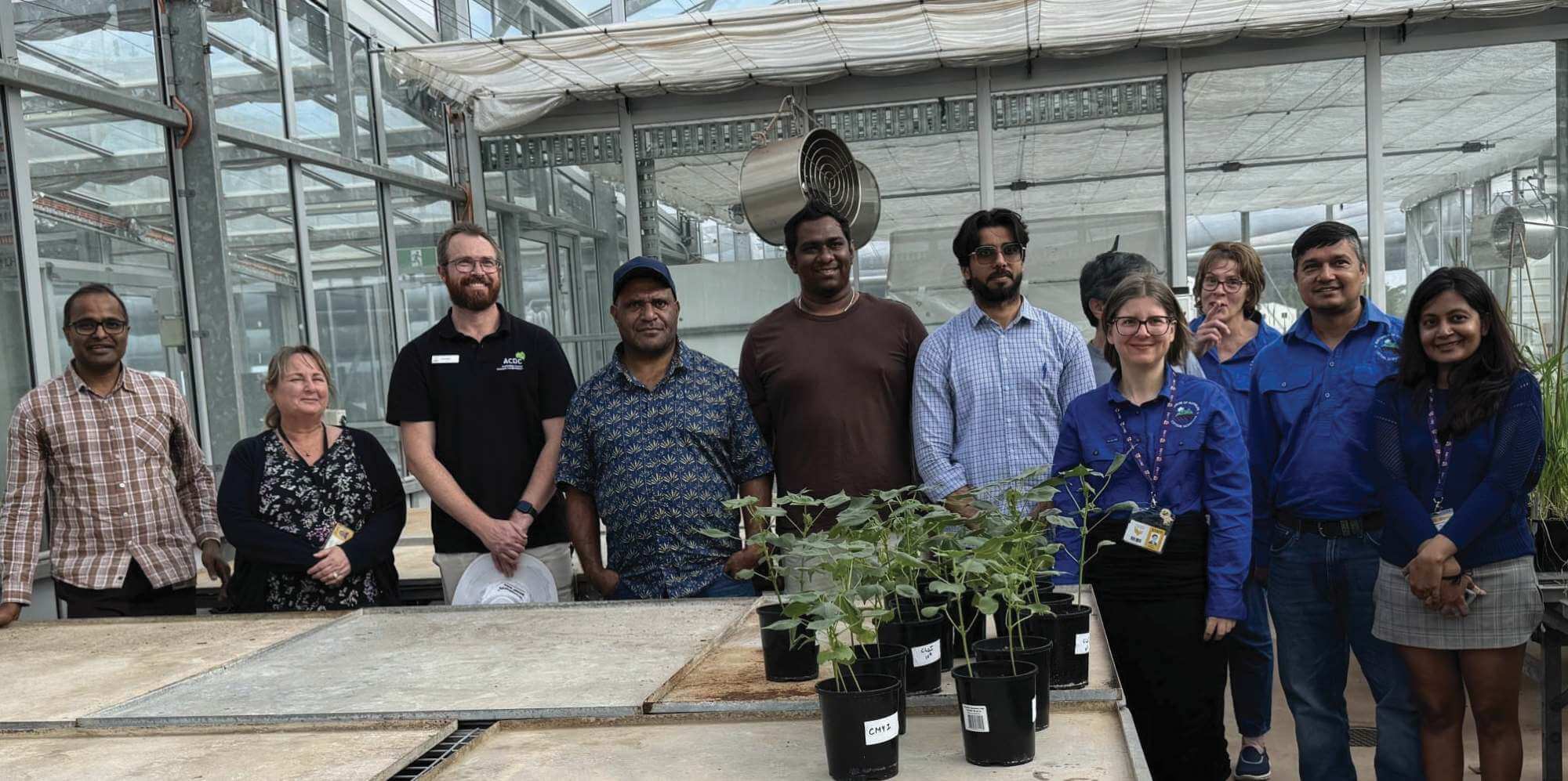 A group of people stand together in a greenhouse behind tables with potted plants, posing for a group photo.