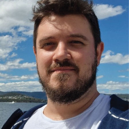A bearded man in a white shirt stands outdoors in front of a body of water, with blue sky and clouds in the background.