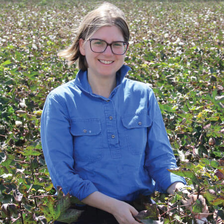 A person wearing glasses and a blue shirt sits in a field of green plants, smiling at the camera.