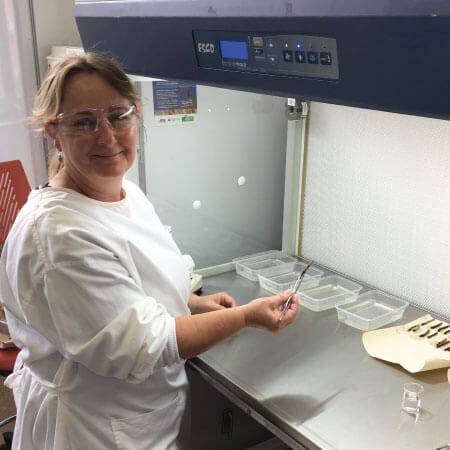 A woman in a lab coat and safety glasses works at a laboratory bench with several clear containers and lab equipment.