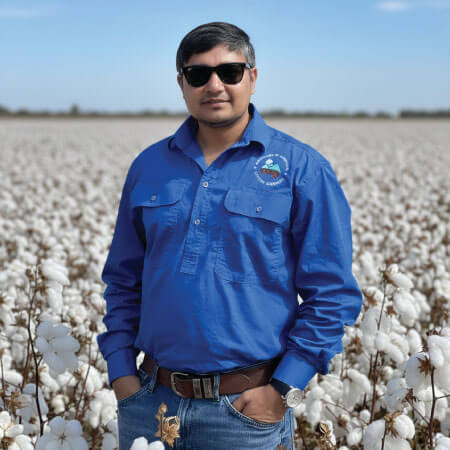 A man wearing sunglasses and a blue shirt stands in a cotton field under a clear sky, with hands in his pockets.
