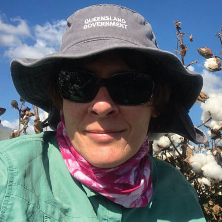 Person wearing a "Queensland Government" hat, sunglasses, and a pink scarf stands in front of cotton plants under a blue sky.