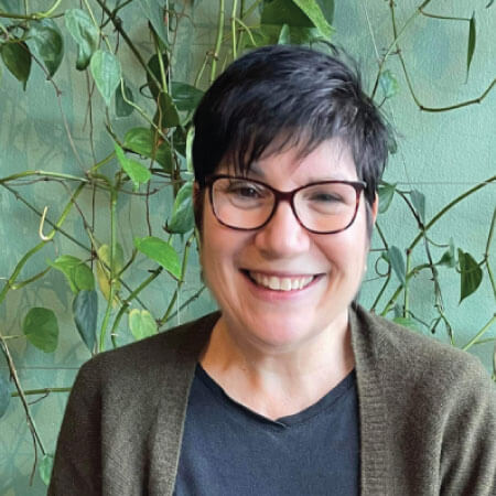 A person with short dark hair and glasses smiling, wearing a dark shirt and brown cardigan, sitting in front of a green wall with leafy plants.