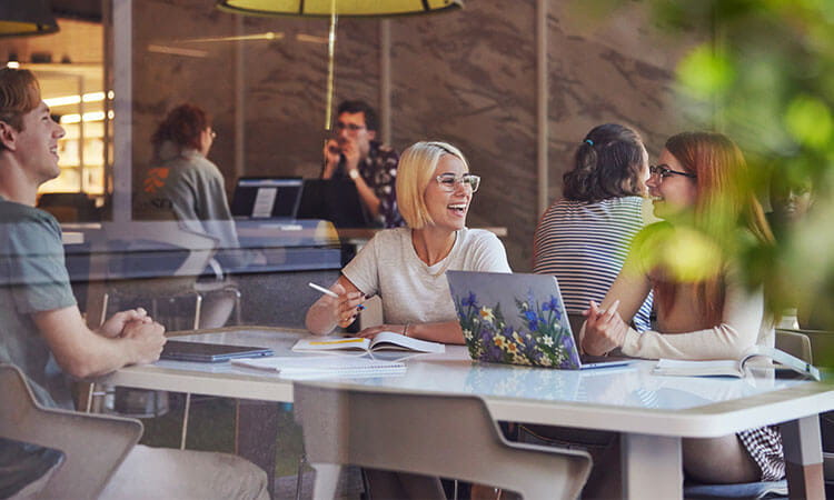 Group of five adults in a casual business meeting at a table in a brightly lit office. One woman is speaking and smiling, others are listening and taking notes.