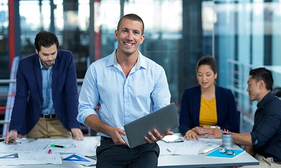 Confident businessman with a laptop standing in an office with colleagues working in the background.