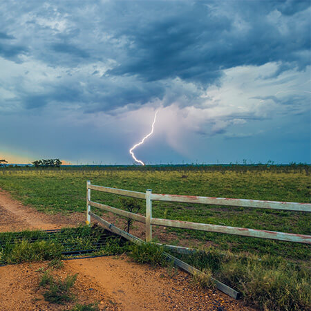 Lightning strikes in the distance over a grassy field under a cloudy sky, with a white fence and dirt path in the foreground.