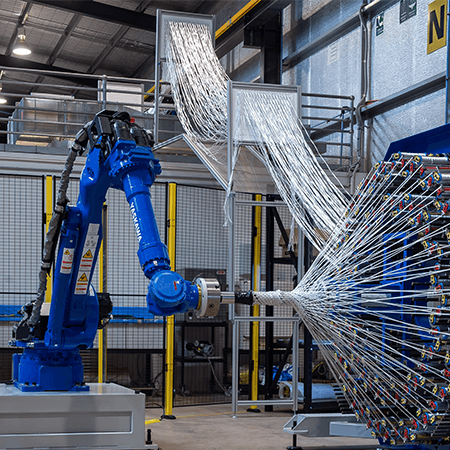 A blue industrial robot arm operates a weaving machine, with numerous white threads extending from a circular component in a factory setting.
