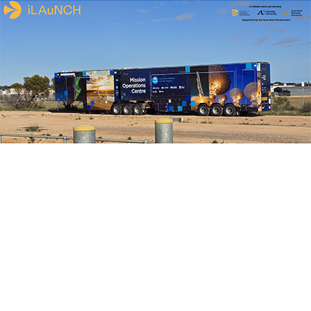 A large blue trailer labeled "Mission Operations Centre" is parked on a dirt lot under a clear sky, with trees and buildings in the background.