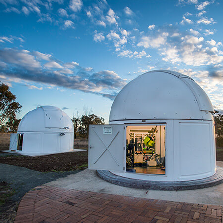 Two white observatory domes with open doors, showing telescopes inside, are set on a paved area under a partly cloudy sky at sunset.