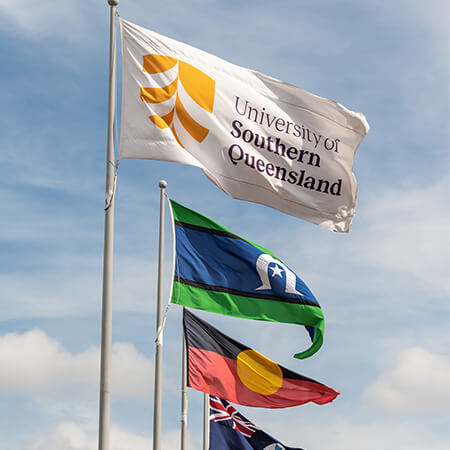 Three flags flying on flagpoles: University of Southern Queensland flag at top, Torres Strait Islander flag in the middle, and Aboriginal Australian flag below. Blue sky background.