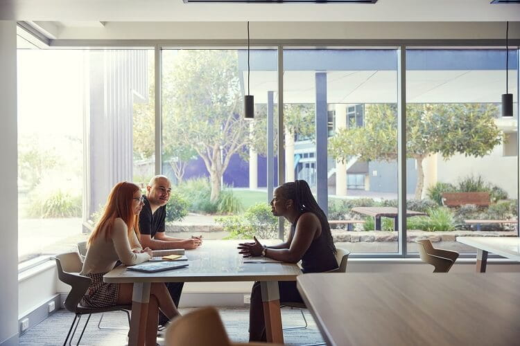 Three people sit at a table in a modern office, engaged in conversation, with large windows and trees visible in the background.