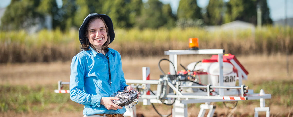 A woman holding a remote control with a drone and agricultural land in the background.