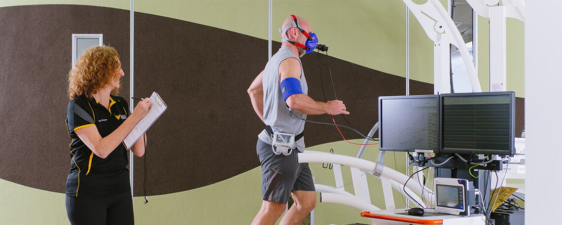 A man with monitoring equipment attached to his body walks on a treadmill while a female technician takes notes next to him.