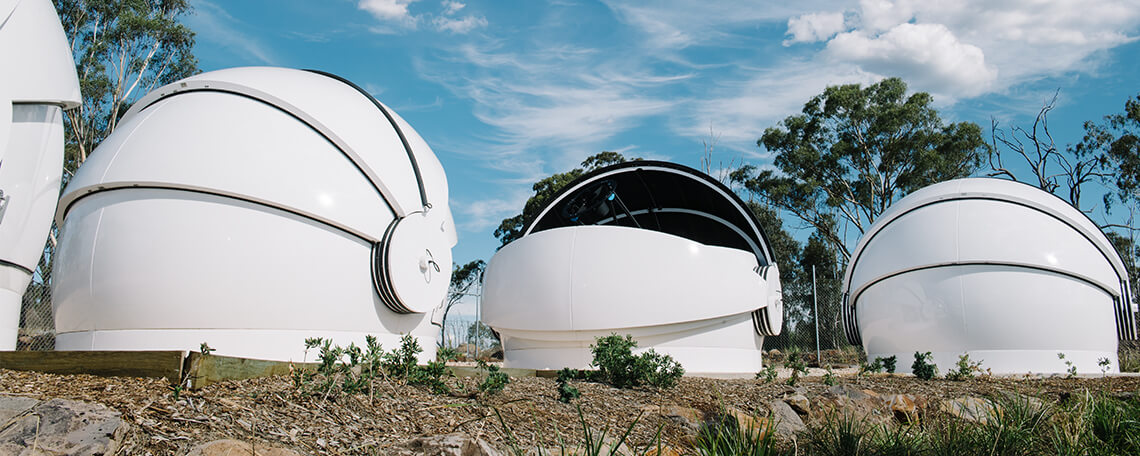 Three white, dome-shaped observatory structures under a blue sky with scattered clouds at UniSQ's Mt Kent Observatory.