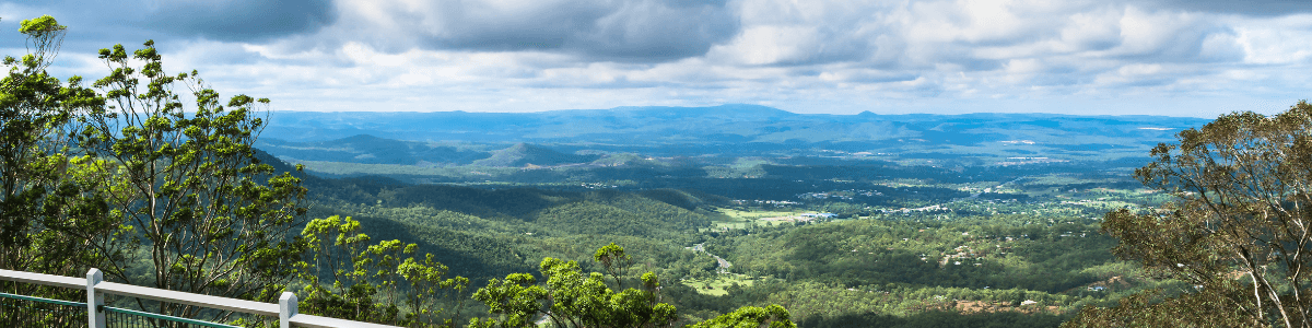 A panoramic view of a lush valley with rolling hills under a cloudy sky, as seen from a viewpoint with a metal railing.