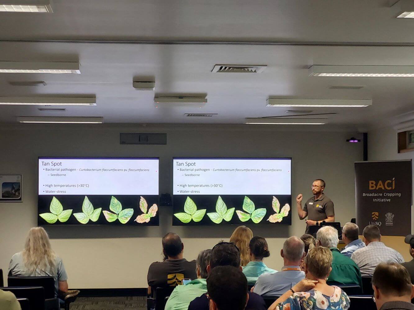 A presenter speaks to an audience about "Tan Spot" in plants, with slides showing plant images and information on temperature and stress factors.