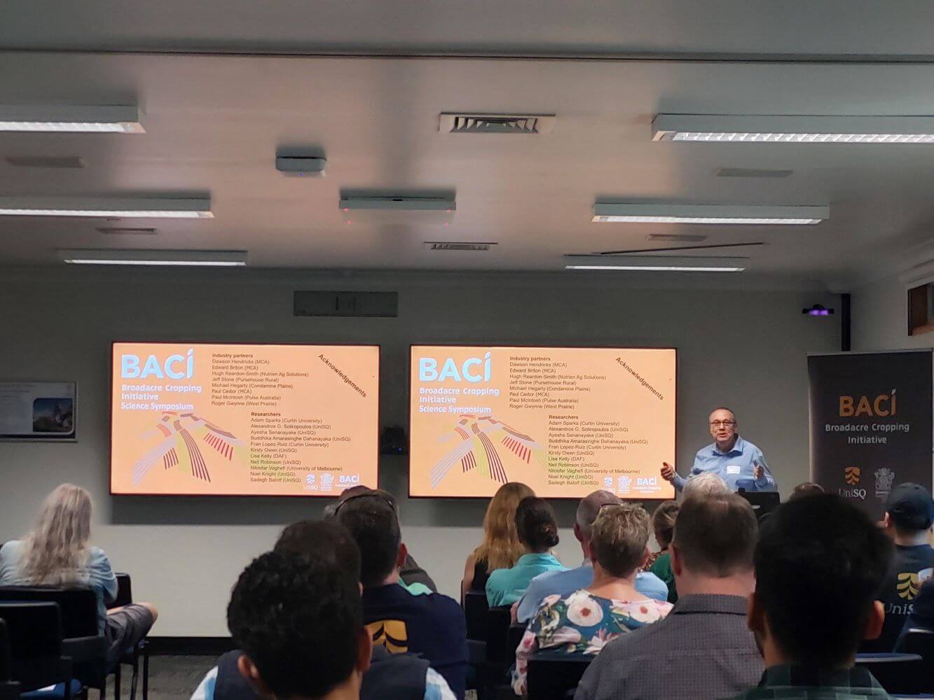 People sitting in an auditorium facing a speaker and two screens displaying a presentation on the "Broadacre Copping Science Initiative" (BACI).