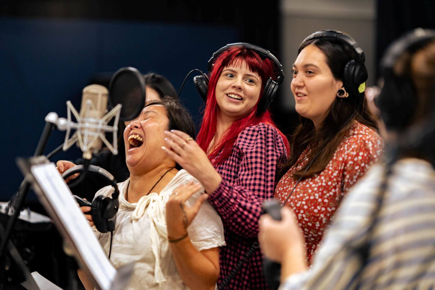 Four women wearing headphones stand in a recording studio, singing and laughing together in front of a microphone.