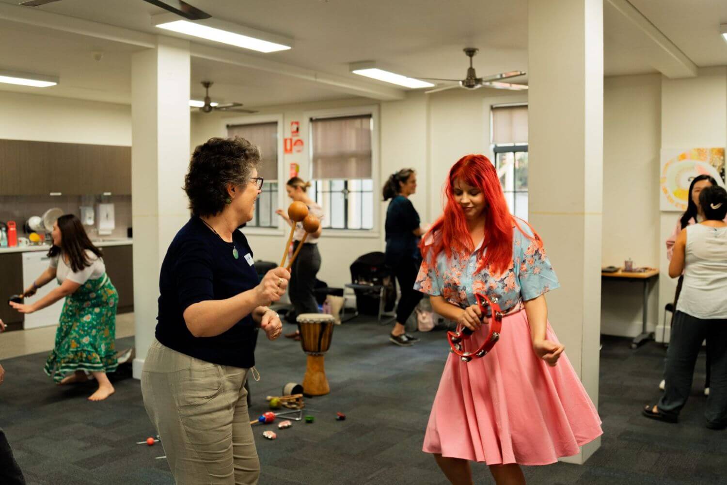 A group of adults plays musical instruments and dances in a room with scattered percussion instruments on the floor.