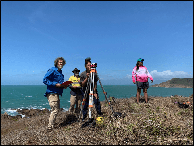 Four people stand on a rocky coastal hillside with surveying equipment under a clear blue sky, facing the ocean.