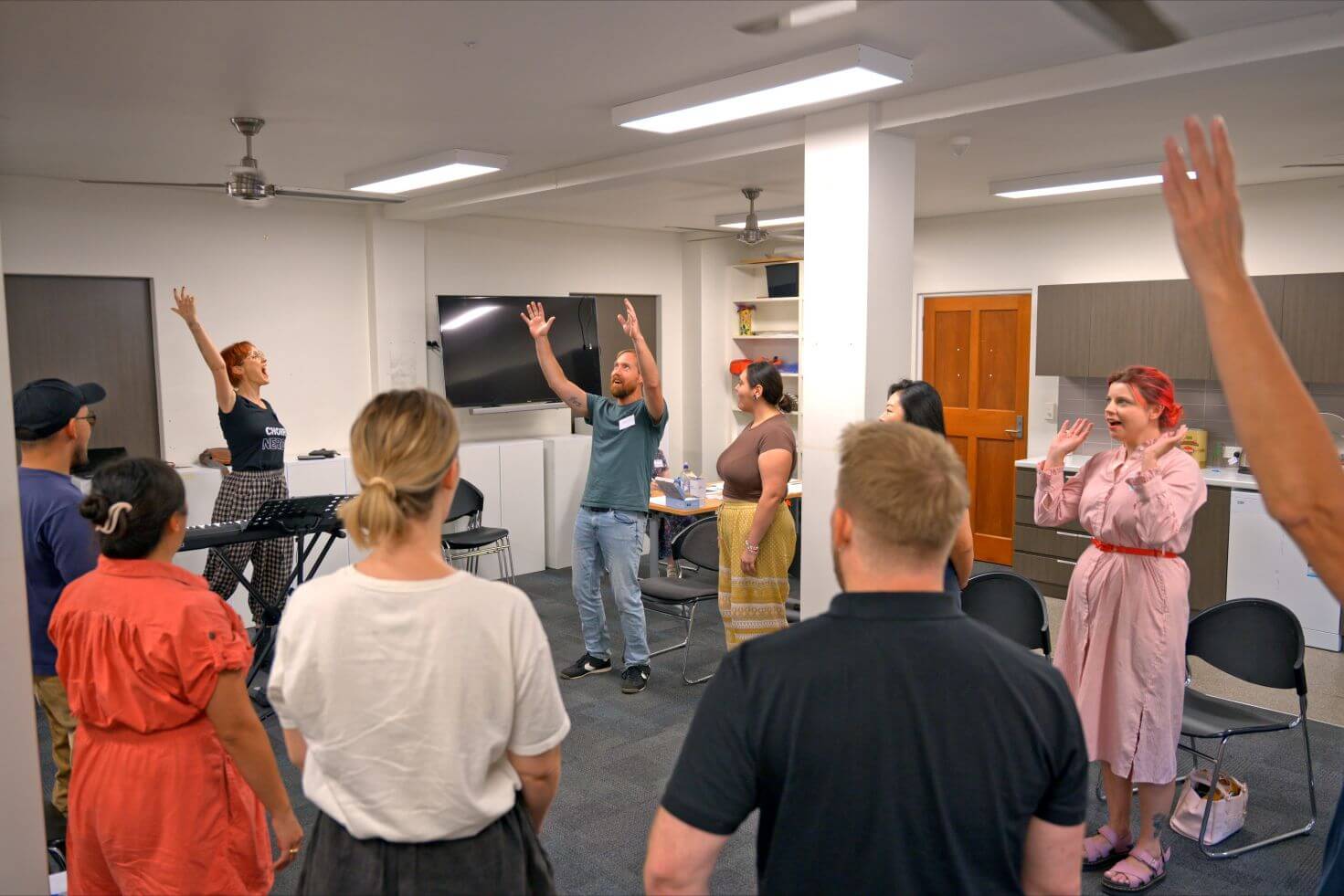 A group of people stand in a circle indoors, raising their arms while participating in an activity led by a woman near a keyboard.